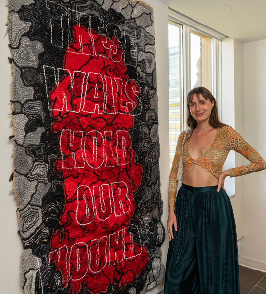 This photo is of student curator Erin Storus standing beside a 6 foot tall red and black textile art work with words embroidered in the centre "These Walls Hold Our Wounds"